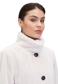 Biały płaszcz z futra sztucznego z wysokim kołnierzem, wyposażony w dwa czarne guziki i miękką, teksturowaną powierzchnię. Zaprojektowany z myślą o ciepłocie i komforcie.