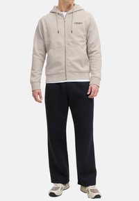 Felpa beige con zip, cordini e tasche frontali, abbinata a pantaloni della tuta neri. Il materiale sembra morbido e dalla texture piacevole.