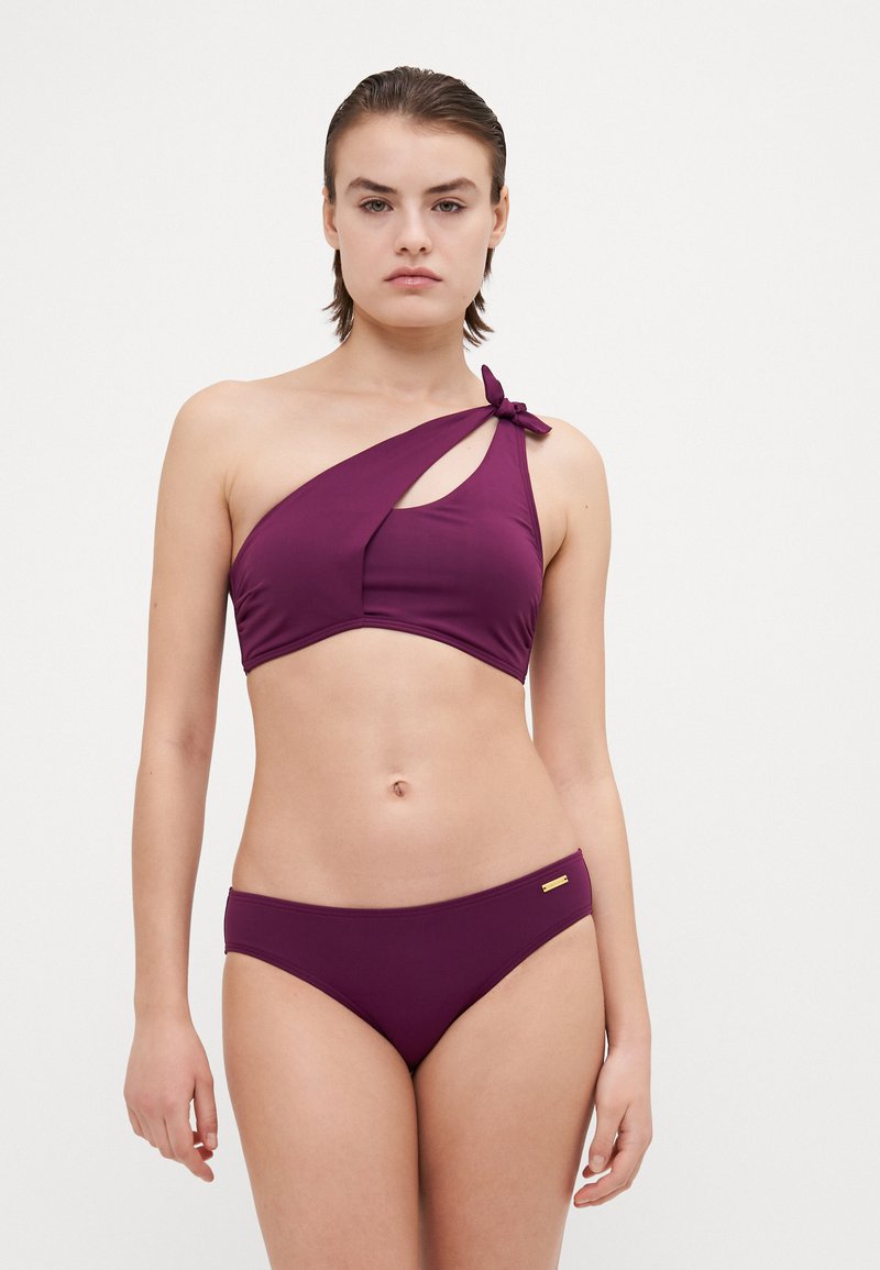 Completo bikini in un intenso viola, con reggiseno a spalla unica con dettaglio nodo e taglio, abbinato a slip a vita bassa. Materiale liscio e elastico.