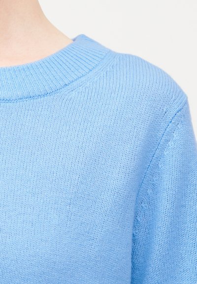 Ανοιχτό μπλε πλεκτό πουλόβερ με ριμπ ύφασμα στο λαιμό και ομαλή υφή υφάσματος στο πάνω μέρος του κορμού και τον ώμο ενός ατόμου.