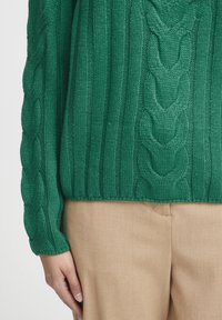 Grüner Strickpullover mit einem Zopfmuster, geripptem Textur und langen Ärmeln, kombiniert mit beigen Hosen.