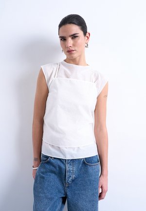 Ung kvinde med mørkt hår samlet bagud, iført hvid ærmeløs top over en hvid skjorte og løse blå jeans, stående foran en ensfarvet hvid baggrund.