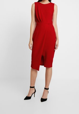 Ærmeløs rød omslagskjole til knæet, båret med sorte hæle med ankelrem, på en stående kvinde mod en ensfarvet baggrund.