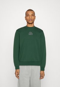 Sudadera verde con cuello redondo, puños y dobladillo acanalados. Presenta un pequeño logo bordado en el pecho. Material de algodón suave.