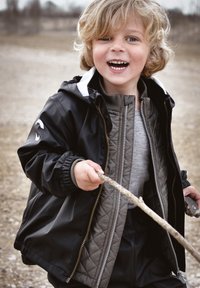 Črna nepremočljiva jakna z belimi poudarki, siva prešita notranja plast, siva majica z dolgimi rokavi, otrok, ki na prostem drži palico.