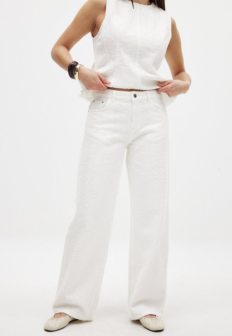 Biely textúrovaný bezrukávový top a široké nohavice, oba s rozstrapkanými okrajmi, spárované so svetlými plochými topánkami.