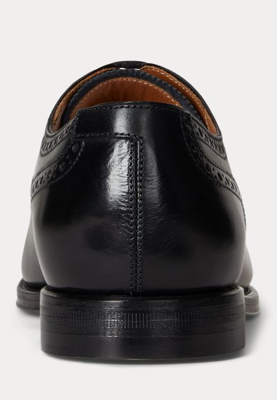 Zapato de vestir de cuero negro con detalles brogue, visto desde atrás, mostrando un acabado pulido y tacón apilado.
