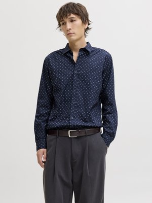 Joven con camisa azul marino de botones con estampado, pantalones gris oscuro y cinturón de cuero marrón, de pie con una mano en el bolsillo.