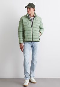 Ljusgrön puffjacka med kviltat mönster, dragkedjestängning och sidofickor. Bärs över en rutig skjorta, ihop med ljusblå jeans och vita sneakers.