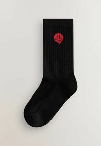 Sorte ribbede sokker med en rund tå, som har et rødt broderet smiley ansigt designet på siden.