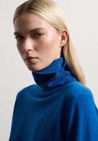 Maglione azzurro a collo alto in maglia, con una texture morbida, che presenta un colletto aderente. Il tessuto appare caldo e accogliente su uno sfondo neutro.