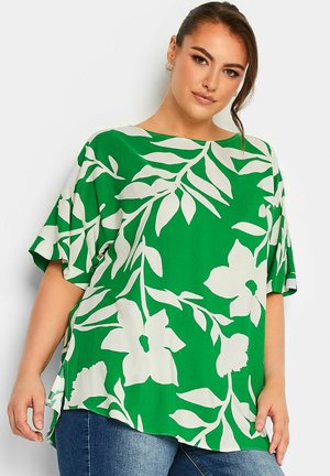 Groene blouse met een groot wit bloemenpatroon, korte mouwen, ruime pasvorm en zijsplitsingen. Lichtgewicht stof met een gladde textuur.
