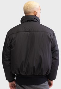 Zwarte pufferjack met een hoge kraag, elastische manchetten en eenGathered taille. De stof heeft een subtiele glans en een gladde textuur.