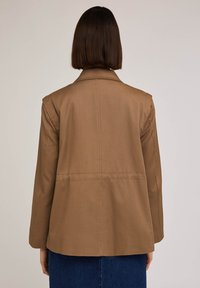 Brązowa marynarka bawełniana z domieszką włókien z wyciętym kołnierzem, długim rękawem i prostym krojem, z plisą z tyłu i minimalnymi detalami przeszycia.