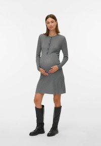 Siva rebrasta pletena obleka z dolgimi rokavi, gumbom spredaj in dolžino do kolen. Manekenka nosi črne škornje do kolen in drži nosečniški trebuh.