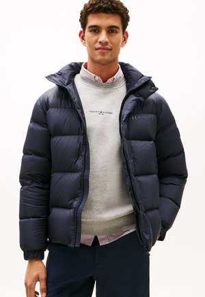 Navy puffer jacket with a zip front and a hood, featuring a quilted design. Worn over a light gray sweater with a logo.