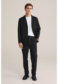 Svart kavaj med enkelknäppt knäppning, kompletterad med matchande byxor. Bärs över en vit t-shirt, har två framfickor och slätt tyg.