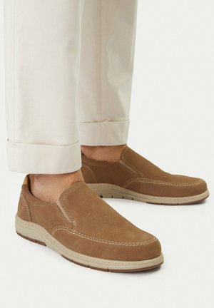 Zapatos de ante marrones sin cordones con costuras beige, usados con pantalones color crema enrollados sobre un fondo blanco.