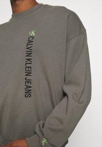 Sudadera gris de algodón con mangas largas; presenta un texto de logo vertical negro y un pequeño logo verde en el puño. Textura suave, corte relajado.