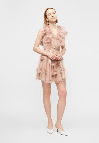 Νεαρή γυναίκα φορώντας ένα ροζ αμάνικο μίνι φόρεμα με βολάν και λουλουδάτα σχέδια, σε συνδυασμό με λευκές μυτερές γόβες, στέκεται μπροστά σε ένα απλό φόντο.