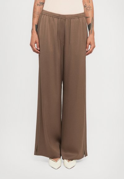 Persone che indossano pantaloni marroni a gamba larga con piccole fessure laterali e scarpe bianche a punta, in piedi contro uno sfondo semplice.