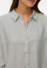 Femme portant une chemise blanche à boutons avec de fines rayures noires verticales et un col, visible des épaules jusqu'au milieu de la poitrine.