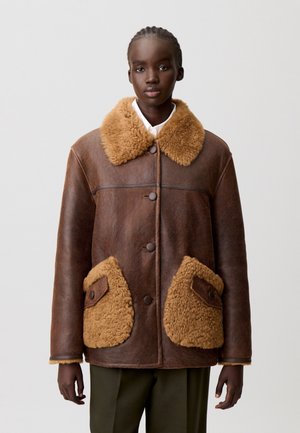 Chaqueta de cuero marrón con un cuello de borreguillo color crema y bolsillos. Presenta botones y costuras visibles, ofreciendo un acabado texturizado.