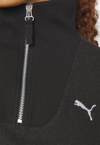Zwarte fleece top met hoge hals, voorzien van een zilveren rits en een wit Puma-logo. Het materiaal heeft een mix van ribbel- en textuurstof.