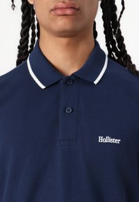 Temno modra polo majica z belim ovratnikom, tri gumbi, in vezenim logotipom "Hollister" na levem prsnem delu. Iz bombažnega materiala.
