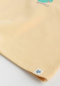 Tela beige con un dobladillo cosido y una pequeña etiqueta blanca con un icono de flor azul cerca del borde.