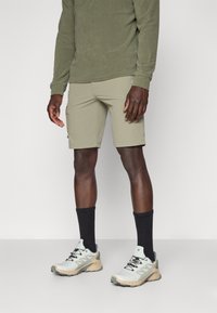 Shorts en khaki à coupe droite avec poches latérales associés à un haut à manches longues vert olive, des chaussettes noires et des chaussures de sport beige clair.