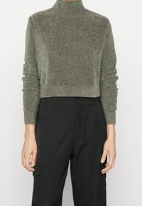 Oliwkowo-zielony krótki sweter dzianinowy z wysokim golfem i dopasowanymi ściągaczami. Połączony z czarnymi spodniami z bocznymi kieszeniami. Gładka faktura.
