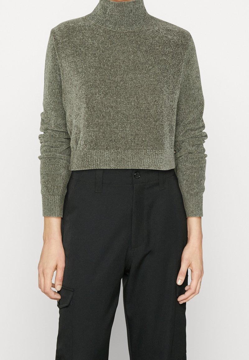 Oliwkowo-zielony krótki sweter dzianinowy z wysokim golfem i dopasowanymi ściągaczami. Połączony z czarnymi spodniami z bocznymi kieszeniami. Gładka faktura.