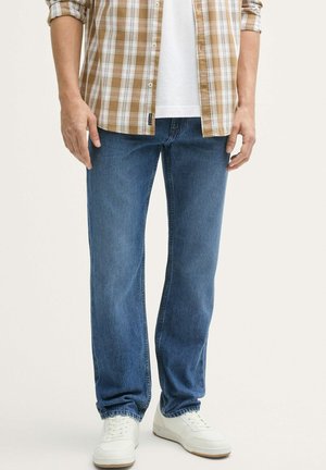 Modri denim jeans s ravnim krojem, kombinirani z rjavim šalom s karirico in belimi supergami. Jeans imajo klasičen zasnova s petimi žepi.
