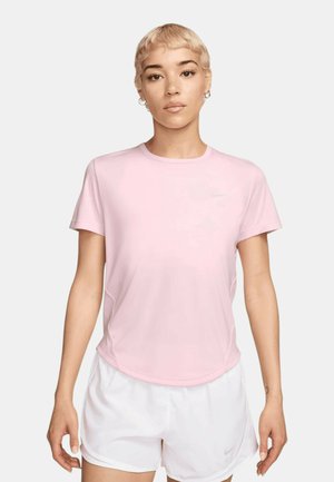 Persoon met kort blond haar die een lichtroze Nike-shirt en witte sportshorts draagt tegen een effen achtergrond.