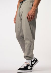 Grijze joggingbroek met elastische tailleband en verstelbaar trekkoord, met zijzakken en contrasterende stiksels, gedragen met zwart-witte sneakers.