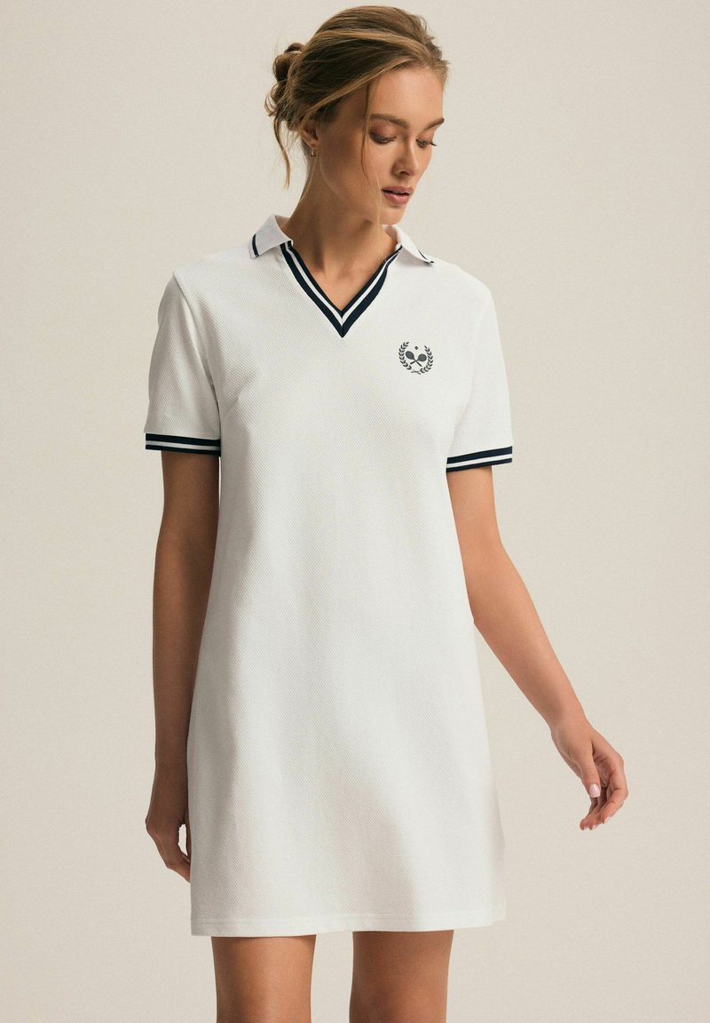 Biała sukienka polo z dekoltem V i krótkimi rękawami. Posiada czarne wykończenia na kołnierzyku i rękawach oraz mały emblemat na piersi. Teksturowana tkanina.