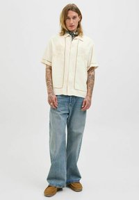 Camisa de manga corta color crema con bordado, combinada con jeans de pierna ancha de color azul claro y calzado marrón. Bufanda atada al cuello.