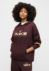 Γυναίκα με σγουρά μαλλιά που φοράει μπουφάν με κουκούλα από τον Nike Athletic Club σε μαρόν χρώμα και αντίστοιχο παντελόνι, στέκεται με τα χέρια στις τσέπες του μπουφάν μπροστά σε απλό φόντο.