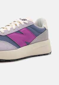 Baskets en suède gris et maille bleue avec un logo "N" violet, des lacets crème et une épaisse semelle beige. Tige texturée avec des accents contrastants.