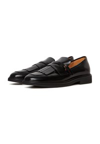Paire de mocassins habillés en cuir noir avec des détails à franges et une bride avec une petite boucle sur fond blanc.