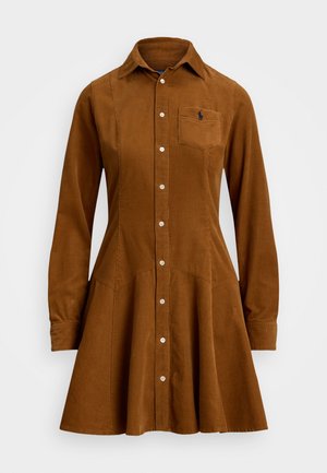 Ruskea corduroy-mekko, jossa on nappikiinnitys, kaulus, pitkät hihat ja rintatasku, jossa on brodeerattu logo. Laskostettu hameosa.