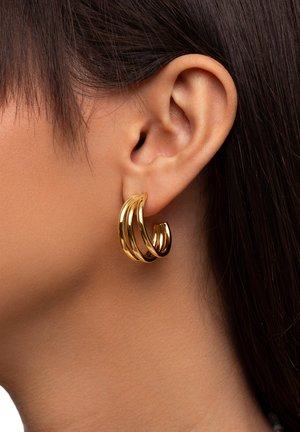 Goldene Creolen mit drei verbundenen Ringen in glänzender Ausführung, die das Licht reflektieren und an das Ohr eines Models lehnen.