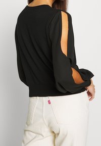 Suéter negro con mangas abullonadas transparentes. Cuenta con un escote redondo y un dobladillo acanalado. Combinado con jeans blancos, mostrando una etiqueta de logo en el bolsillo trasero.