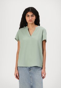 Blusa verde chiaro a maniche corte in un tessuto liscio con scollo a V e una vestibilità rilassata, abbinata a una gonna di jeans.