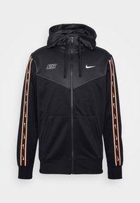 Svart zip-up hoodie med hög krage, med vita Nike-logotyper, orange och vita sidremsor, samt framfickor. Mjuk tygtextur.