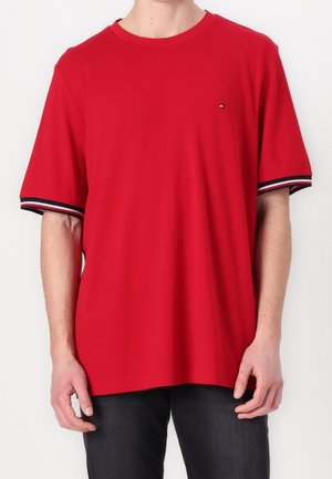 Homme portant un t-shirt rouge uni à manches courtes avec des poignets rayés noir, blanc et rouge et un petit logo sur la poitrine gauche.