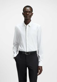 Camicia bianca a maniche lunghe con colletto strutturato, caratterizzata da un bottone nascosto e dettagli di cucitura a contrasto, abbinata a pantaloni neri slim-fit.