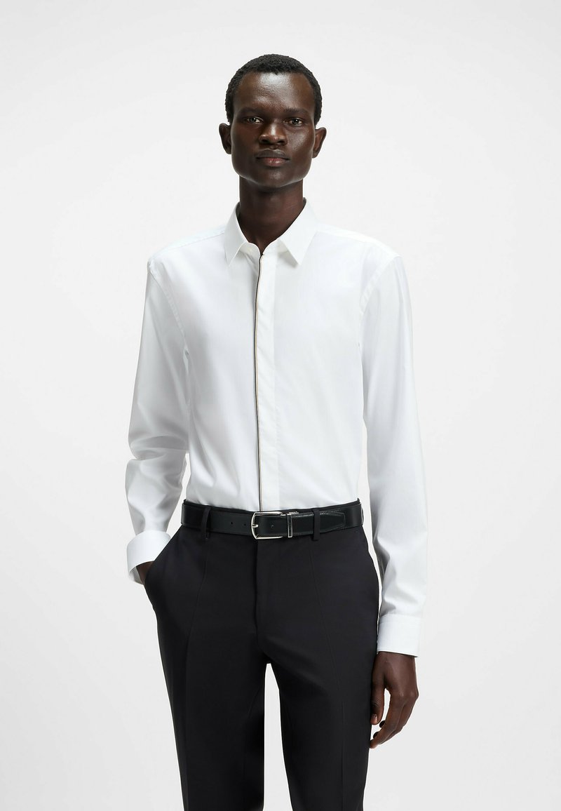 Camicia bianca a maniche lunghe con colletto strutturato, caratterizzata da un bottone nascosto e dettagli di cucitura a contrasto, abbinata a pantaloni neri slim-fit.