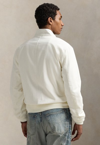 Vyras su trumpais garbanotais plaukais, žiūrintis iš šono, dėvi baltą striukę ir išblukusius mėlynus džinsus prieš vienos spalvos smėlio spalvos foną.